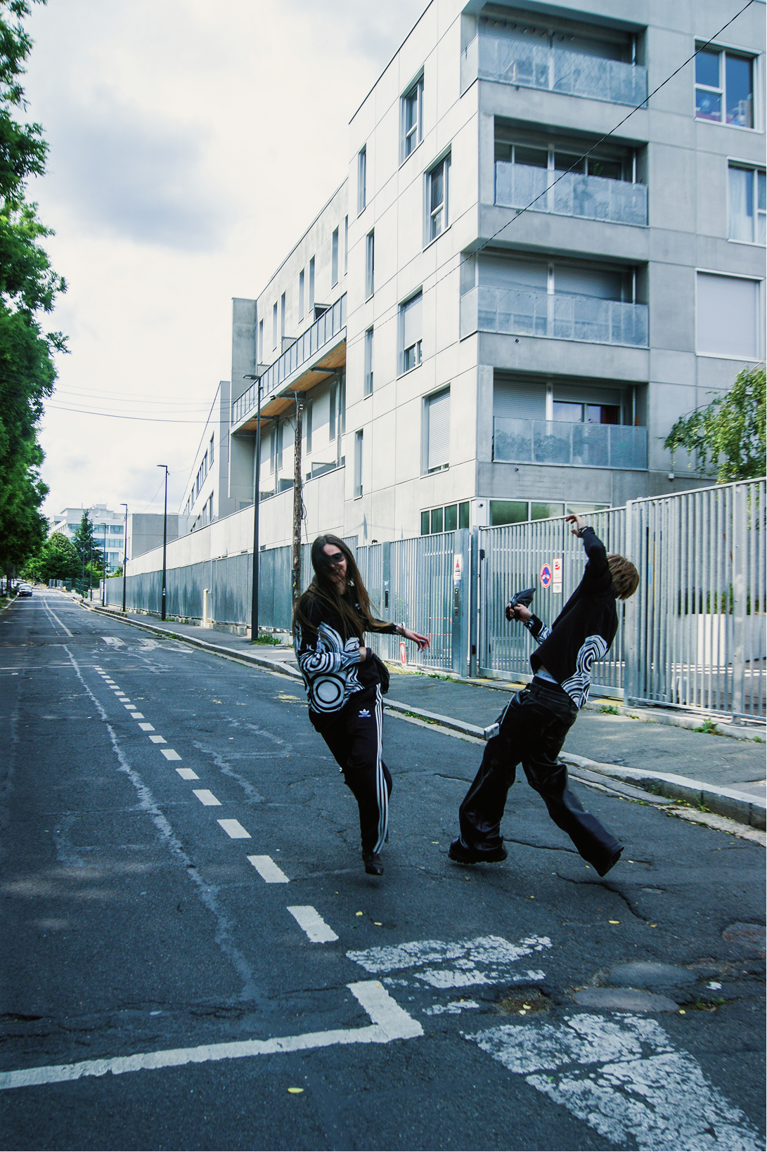 Deux personnes marchant et dansant au milieu d’une rue déserte bordée d’immeubles modernes, habillées de t-shirts noirs à grands motifs graphiques blancs, ambiance streetwear urbaine.